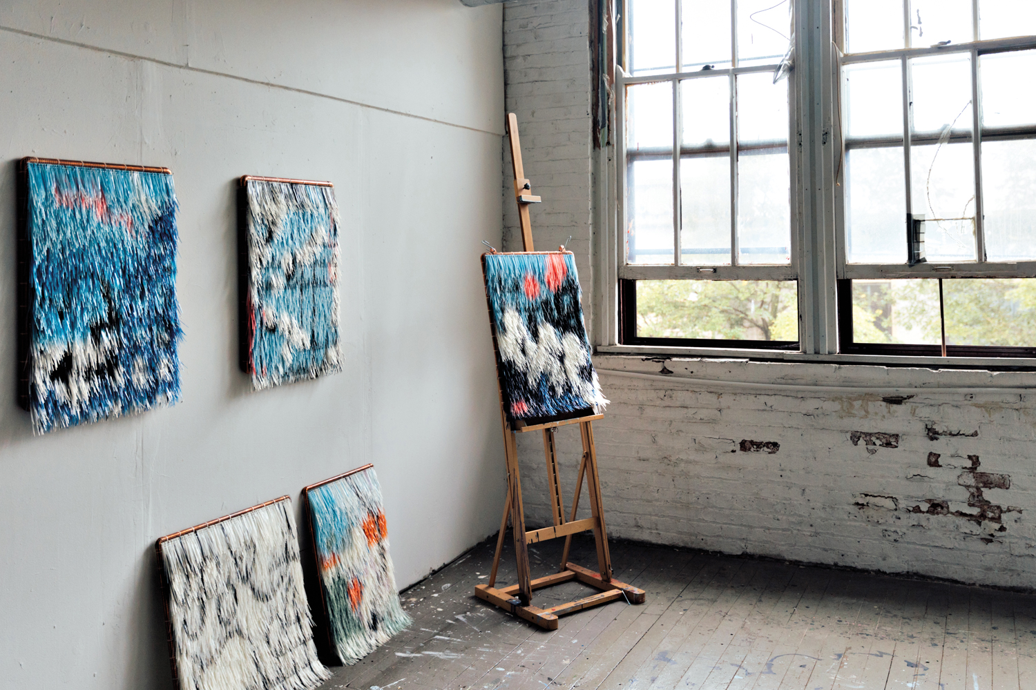 Five fringed artworks- two hanging on a wall, two leaning against a wall and one on an easel