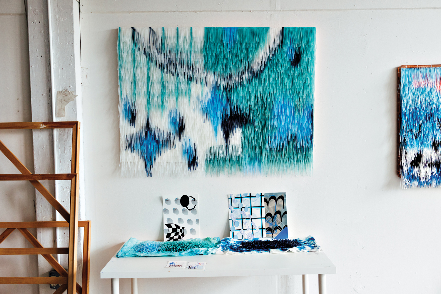 Fringed artworks hang on a studio wall with drawings on a table beneath