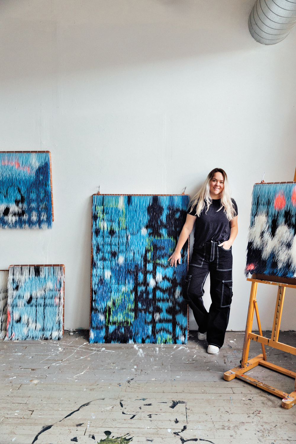 Sarah Leuchtner stands in studio in front of fringed artworks in shades of blue, green and pink