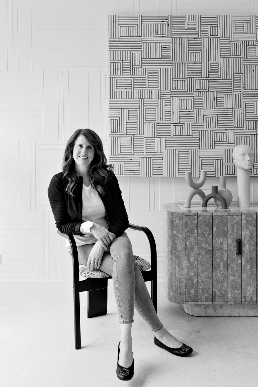 Black-and-white portrait shot of designer Jen Talbot sitting in front of artwork and a cabinet.
