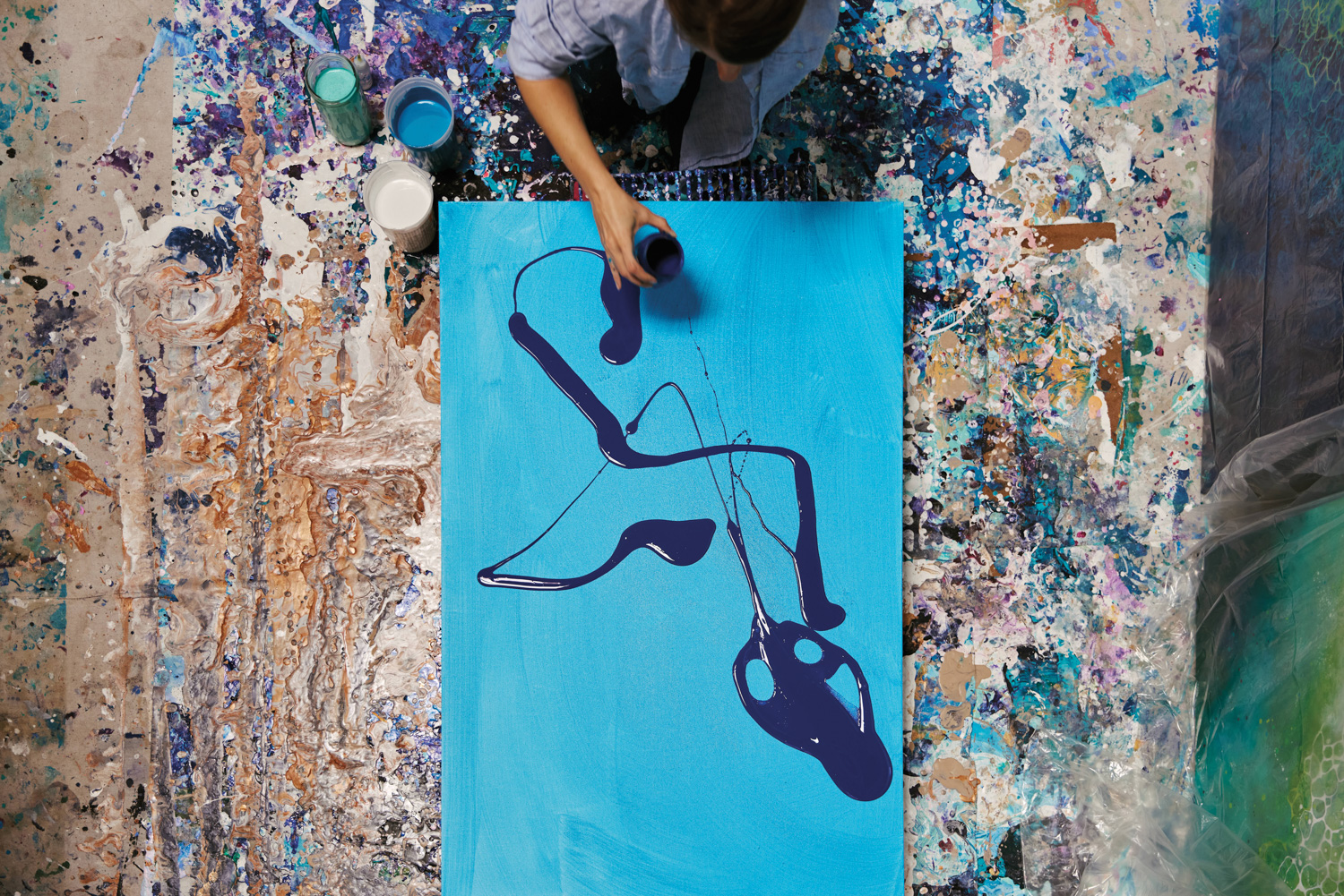 an artist pouring navy paint in swirls over a canvas lying atop paint splattered floors