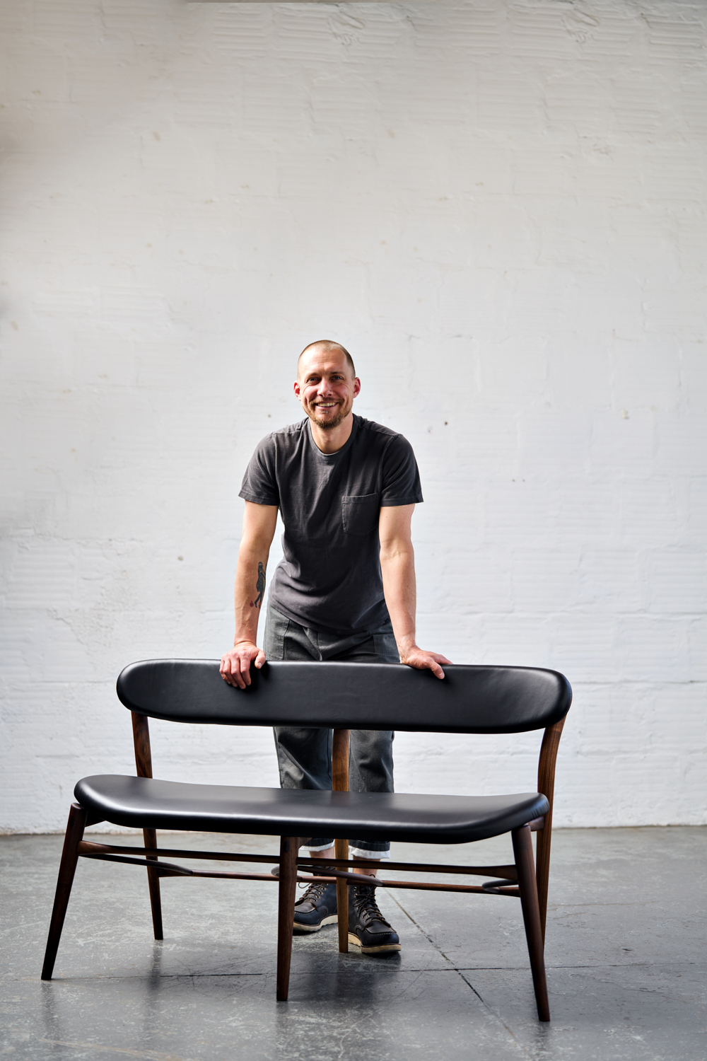 Woodworker Justin Nelson leaning against a dark wood bench with black upholstered seat and back cushions.
