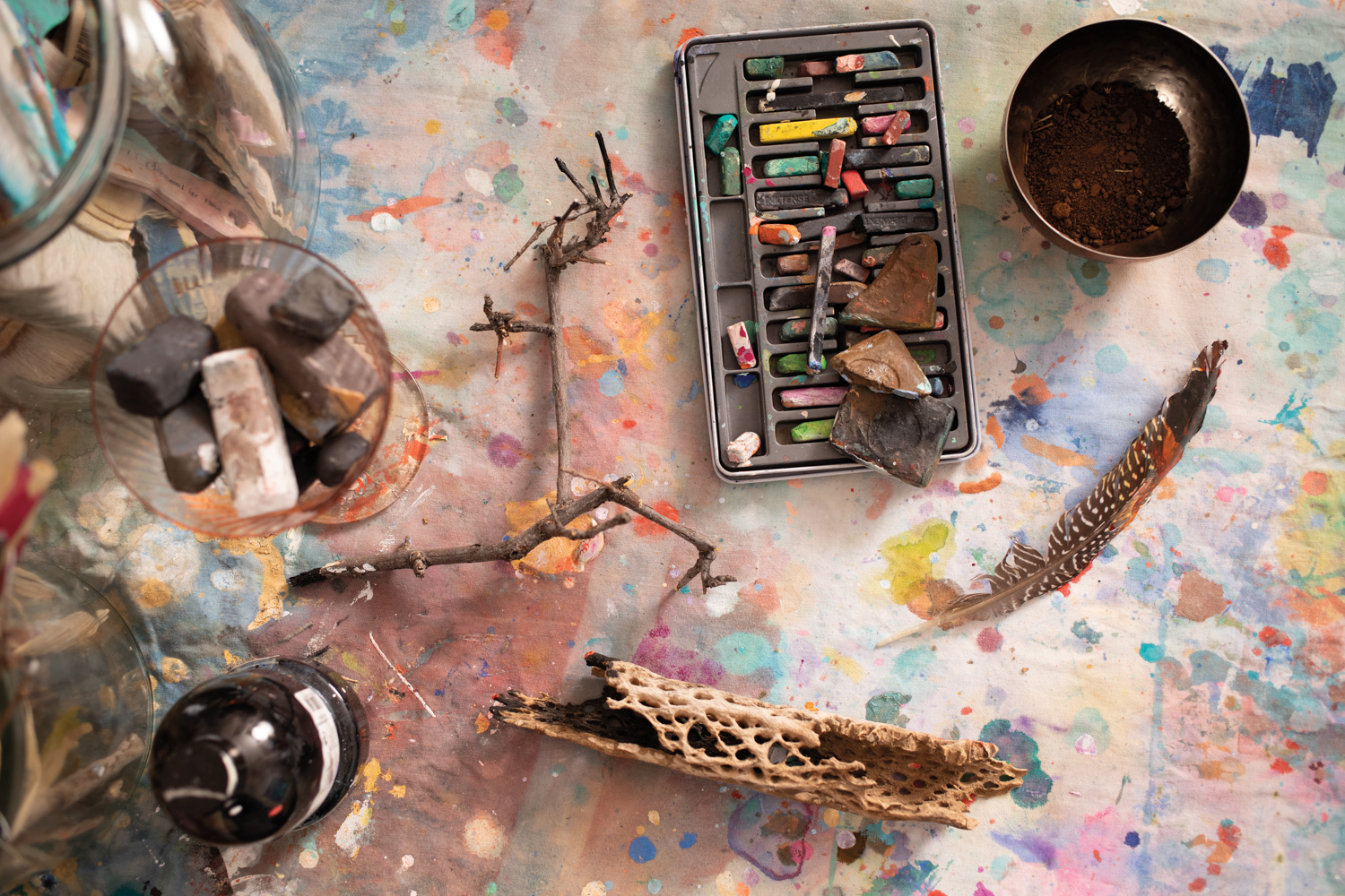 A collection of pastels, a bowl of dirt, driftwood and a feather sit on a paint-splattered table.