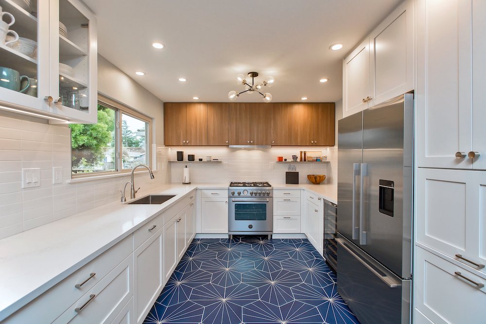 A kitchen featuring a modern design with white lower cabinets contrasting against wood upper cabinets. The blue tile floor adds a vibrant touch to the space. Stainless steel appliances seamlessly blend in, contributing to the contemporary aesthetic of the kitchen.