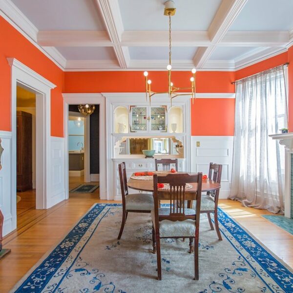 a vibrant dining room with warm orange walls providing a welcoming atmosphere. A sturdy wood table sits atop a blue area rug, anchoring the space with a pop of contrasting color. The white ceilings enhance the room's brightness, creating a visually pleasing and cozy dining environment.