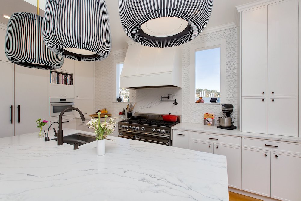 A bright, contemporary kitchen with a cohesive white color scheme, including white cabinets, countertops, and a range. The space is illuminated by stylish black pendant lighting fixtures, creating a striking contrast and adding a touch of modern elegance.