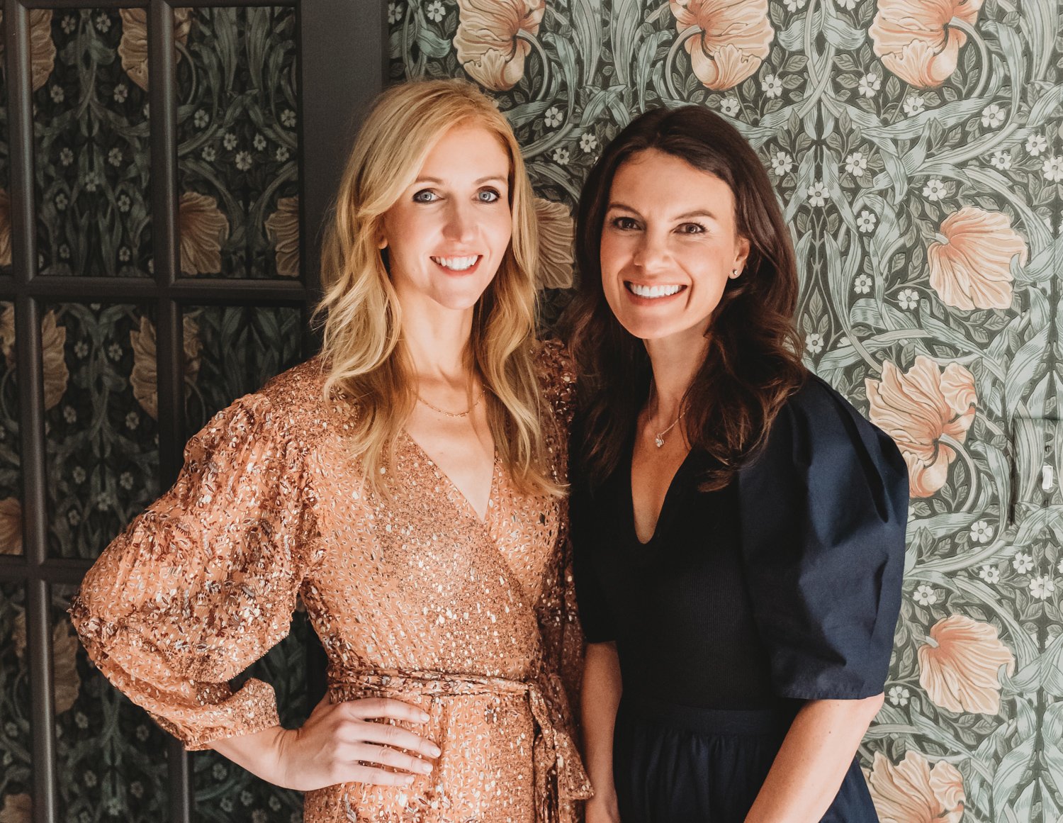 Designers Renee DiSanto and Christina Samatas posing against a floral patterned wallpaper