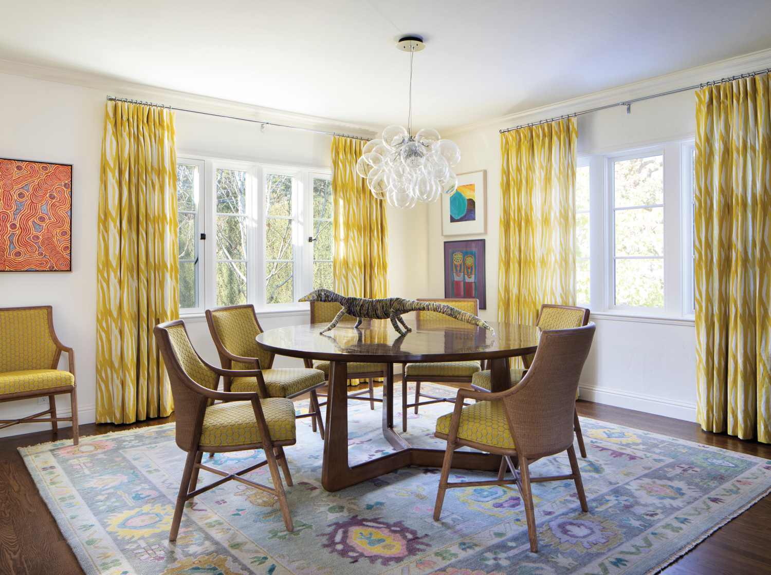 Dining area with bubble-like chandelier over a round table surrounded by yellow upholstered chairs and set against windows flanked by yellow drapes.