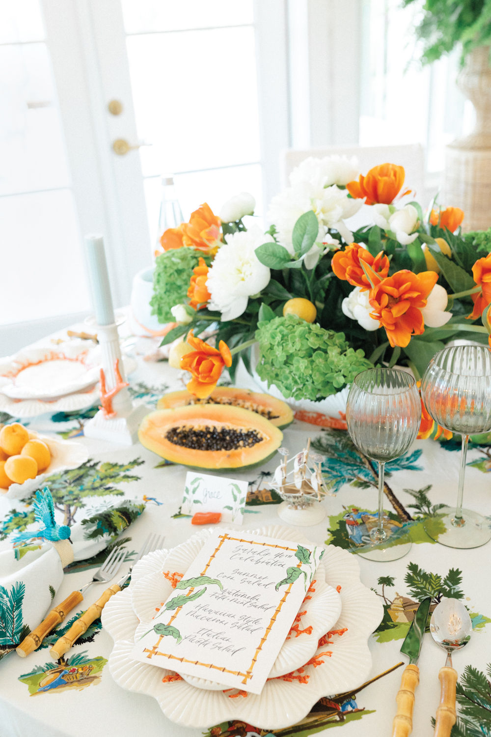Styled tabletop place settings with bamboo-handled utensils and a white, green and orange floral arrangement next to papaya halves.