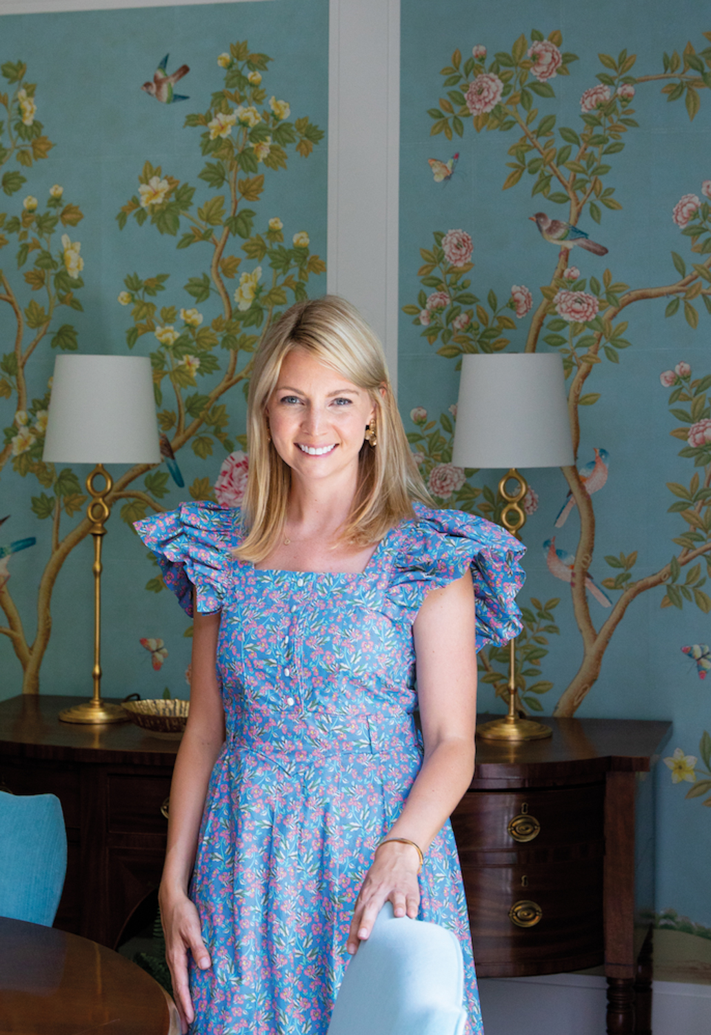Becky Nielsen Filipski stands in a floral dress in a dining space with blue Gracie-like wallpaper