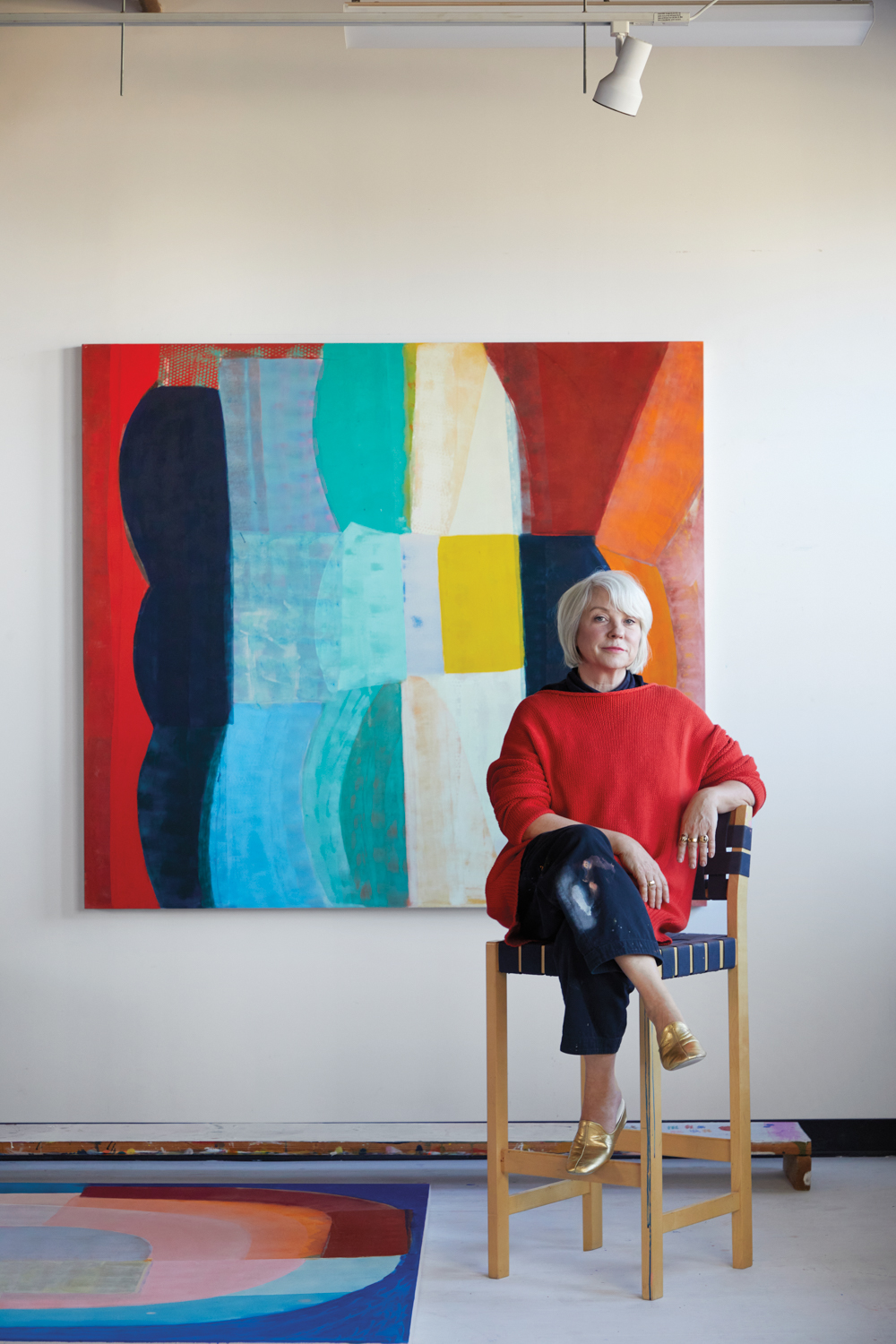woman in red sweater sitting on a stool in front of a large colorful painting