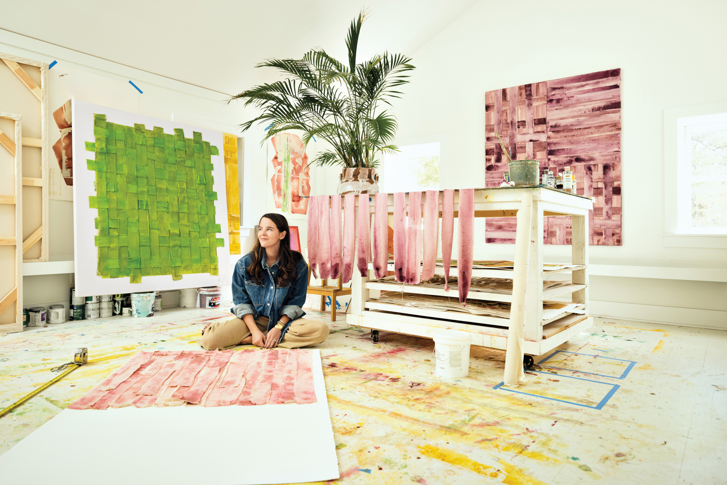 Sitting woman gently smiling within a white room surrounded by colorful artwork and a lush palm tree