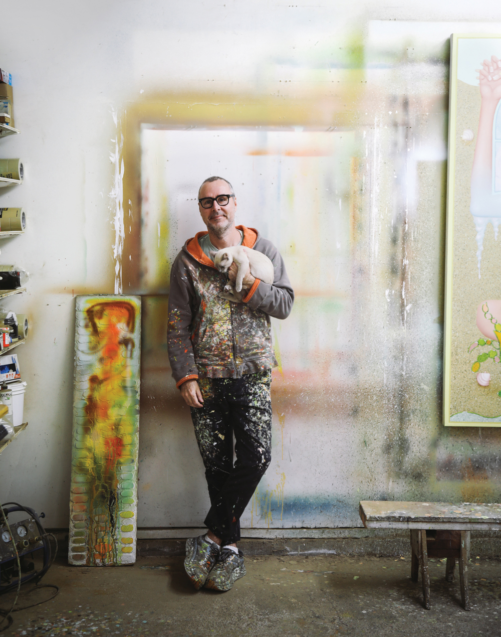 man in paint splatted clothing and glasses holding cat while leaning against a paint covered wall near a low workbench