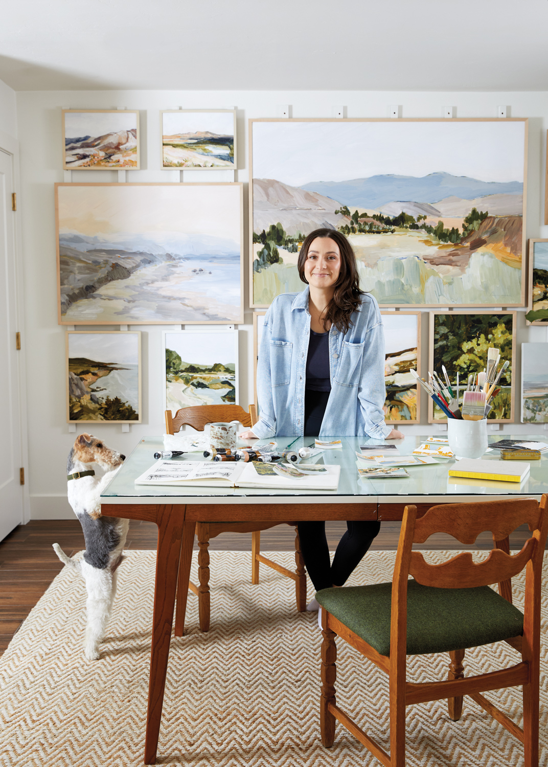 a woman in a blue shirt stands at an art table with landscape paintings behind her and a dog next to her