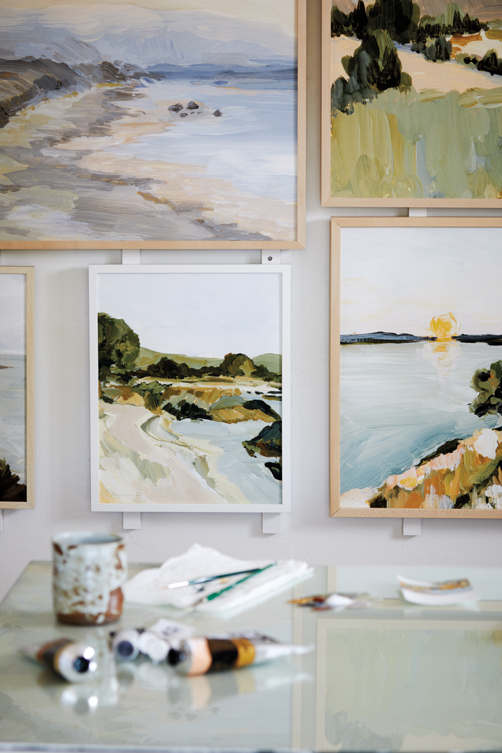 gallery wall of acrylic landscape paintings in muted greens, blues and yellows