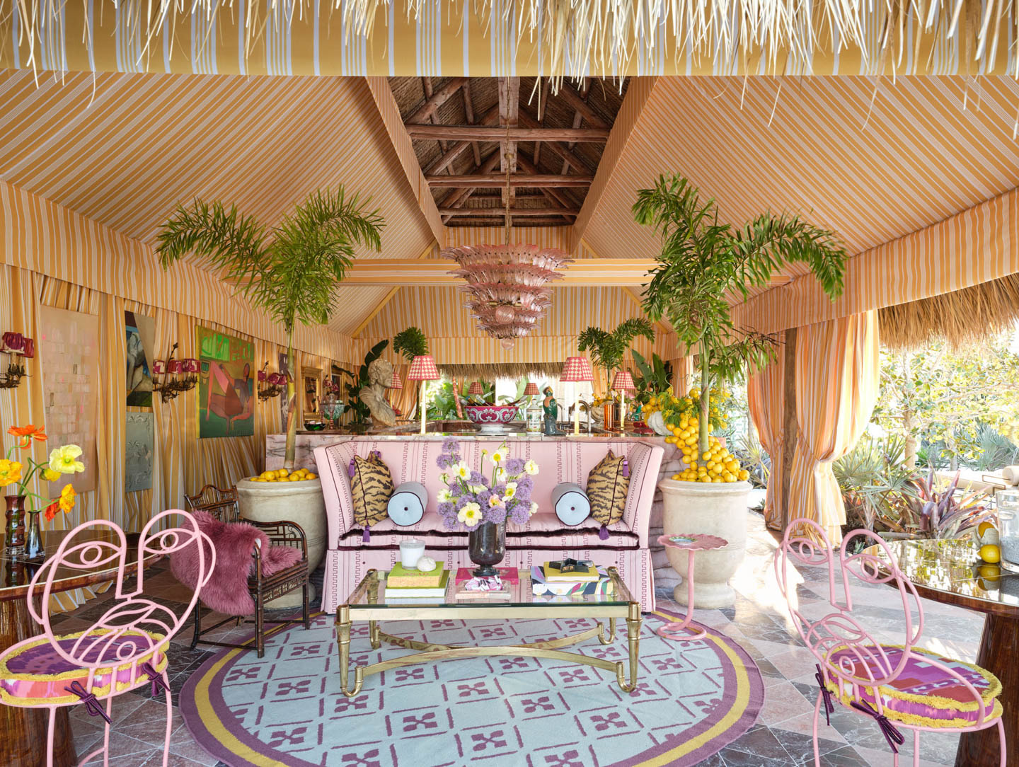 Tropical-themed room with pink and yellow stripes, lush plants, and wicker furniture. Pink chairs, floral cushions, and playful decor set a vibrant, cheerful tone.