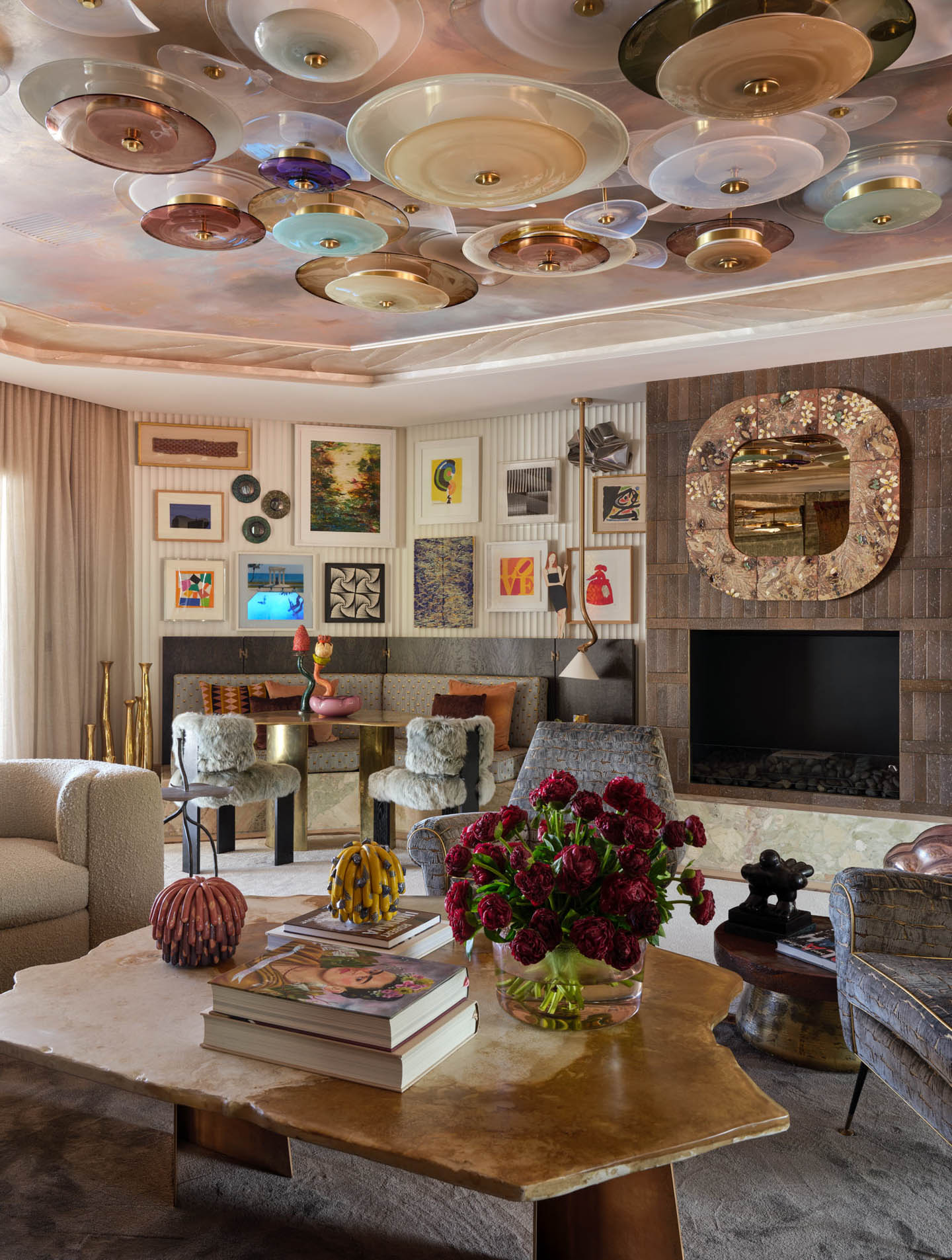 Eclectic living room with a vibrant ceiling art installation. Walls feature diverse artwork. Cozy chairs, a plush sofa, and a rustic table with floral decor create a warm, artistic atmosphere.