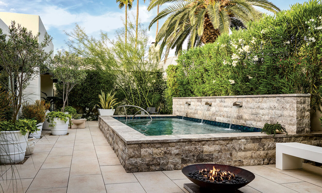 Luxurious patio with a square pool featuring cascading water, surrounded by lush greenery and potted plants. A fire pit and palm trees add warmth.