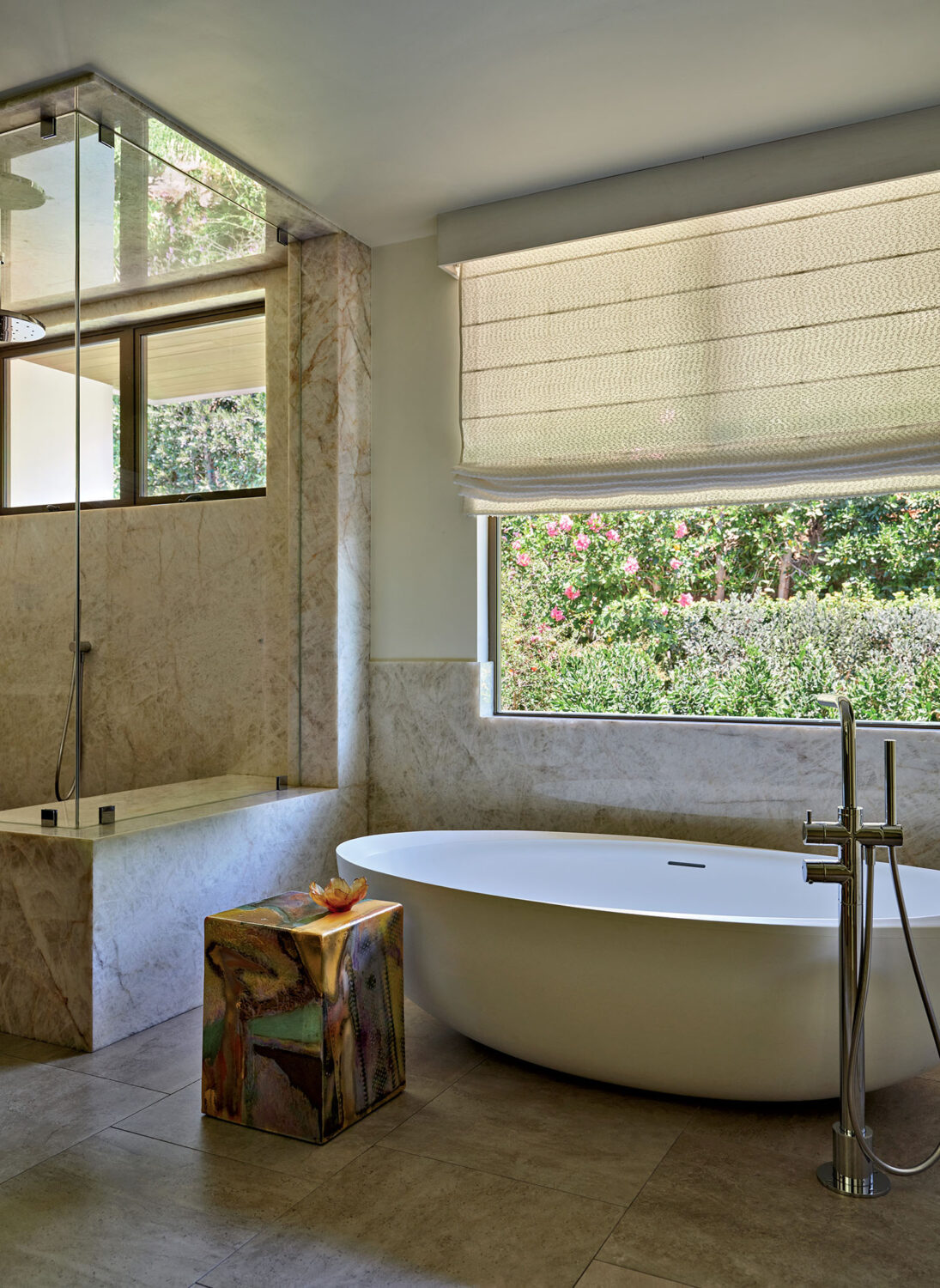 Modern bathroom with a large white oval tub on gray tiles next to a colorful cube table with a flower. Large window with garden view enhances tranquility.