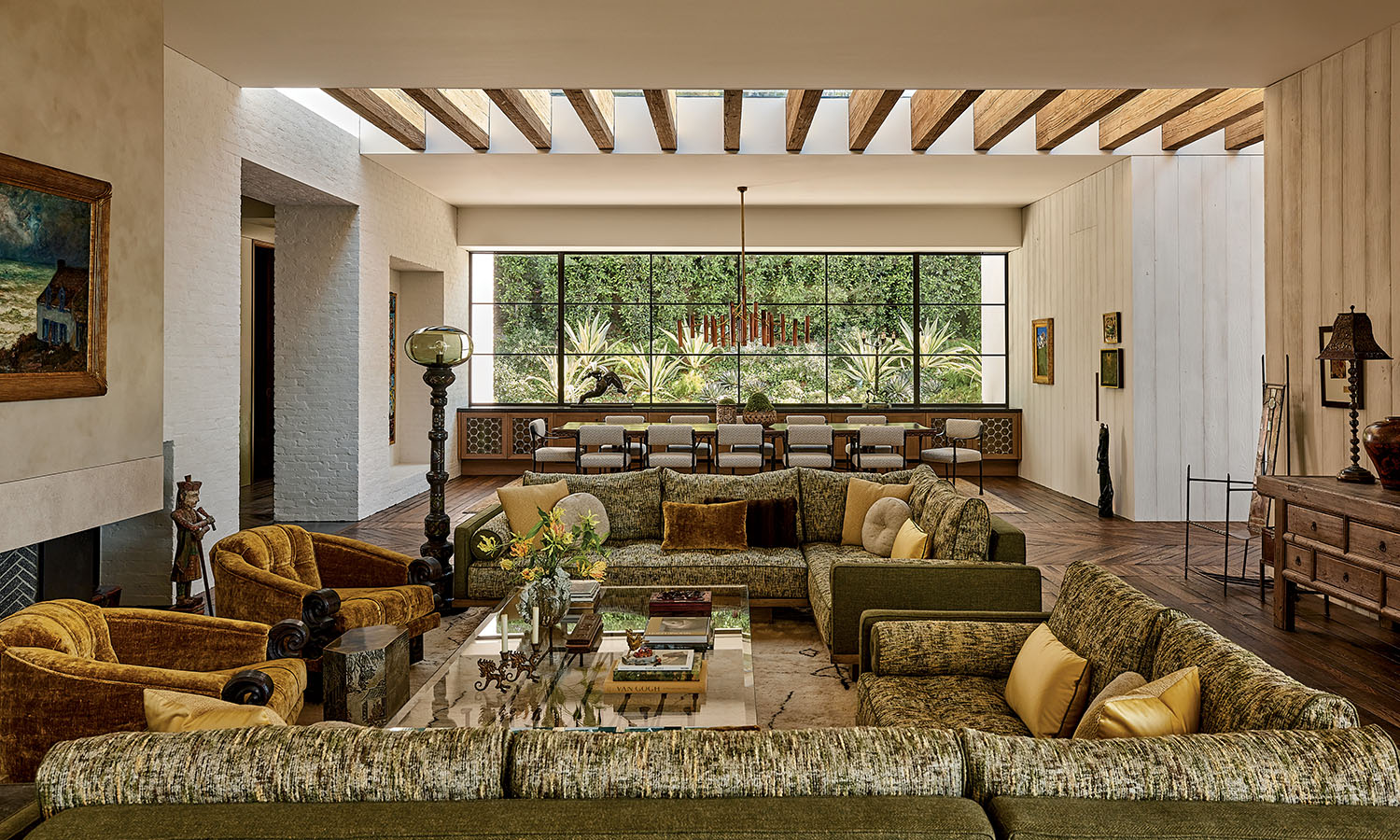 Modern living room with large windows revealing lush greenery. Features a cozy couch with cushions, round dining table, chairs, and stylish pendant light. Bright and serene ambiance.