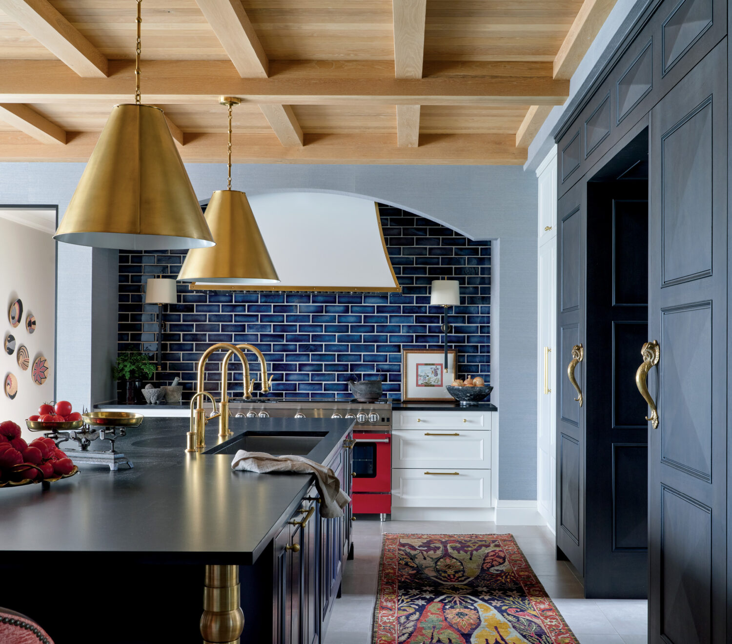 Spacious kitchen with blue tiled backsplash, red oven, and gold fixtures. Featuring a dark island, brass lamps, fruit bowl, and a colorful rug. Elegant and warm.