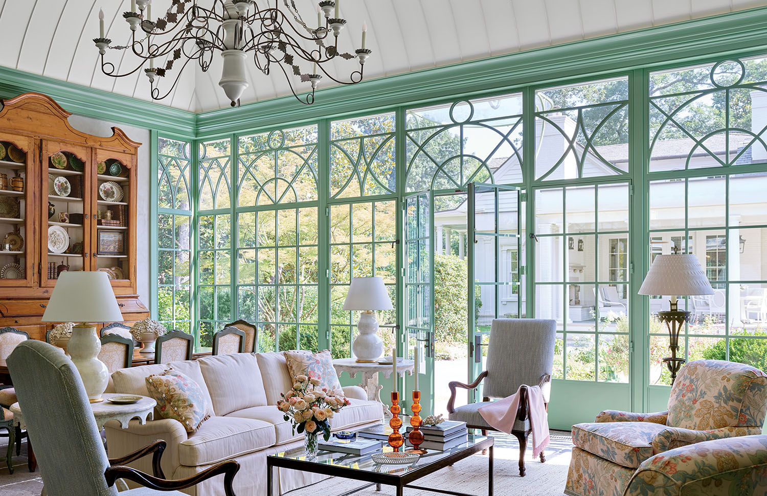 Elegant sunroom with large, green-framed windows, plush sofas, floral armchairs, and a vintage chandelier. Light-filled, cozy, and inviting atmosphere.