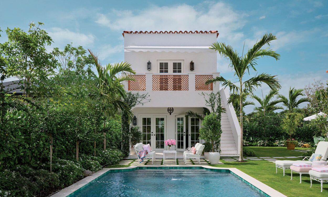 Elegant villa with white facade and terracotta tiles, lush garden, palm trees, and a serene blue pool. Cozy chairs surround the tropical oasis.