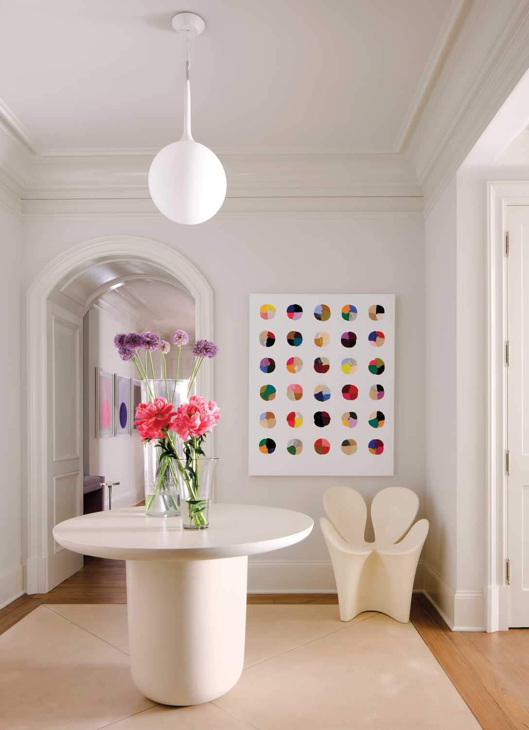 A minimalist room features a round table with vibrant pink and purple flowers in a vase. A colorful dot painting is on the wall, under a modern white pendant light.