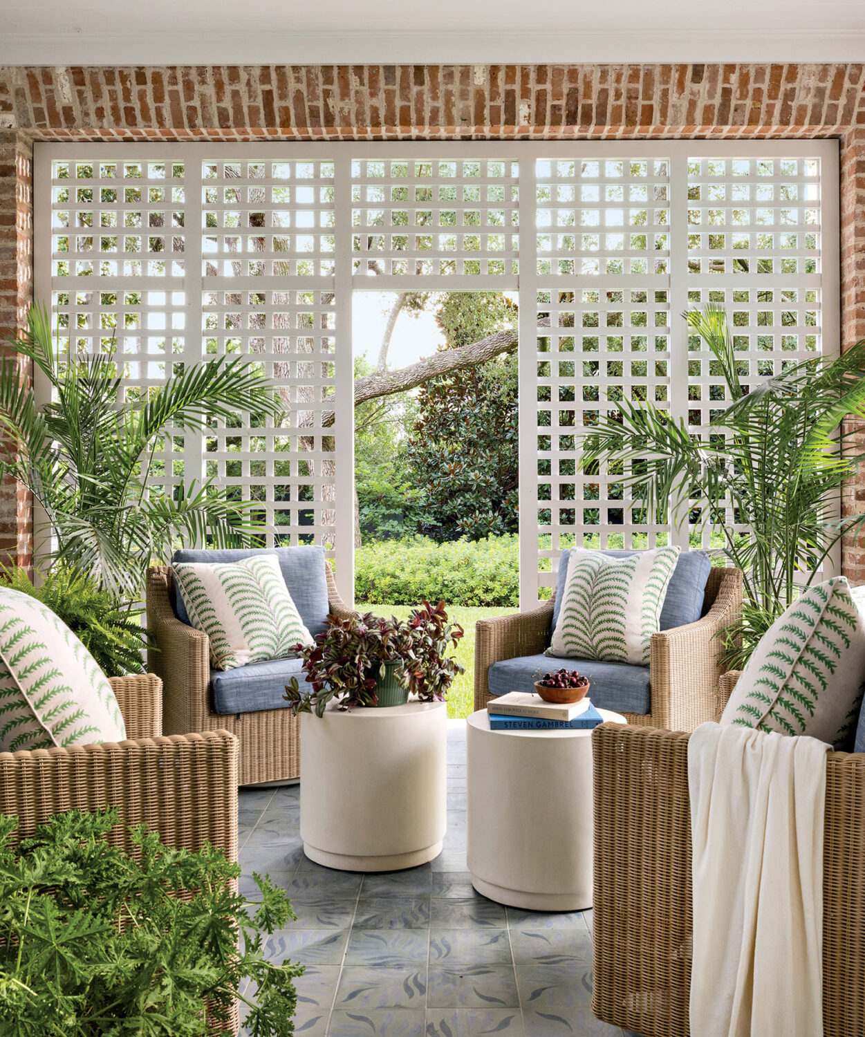Cozy porch with wicker chairs, blue cushions, and leaf-patterned pillows. White lattice wall, lush greenery outside, and ceiling fan overhead. Calm and inviting.