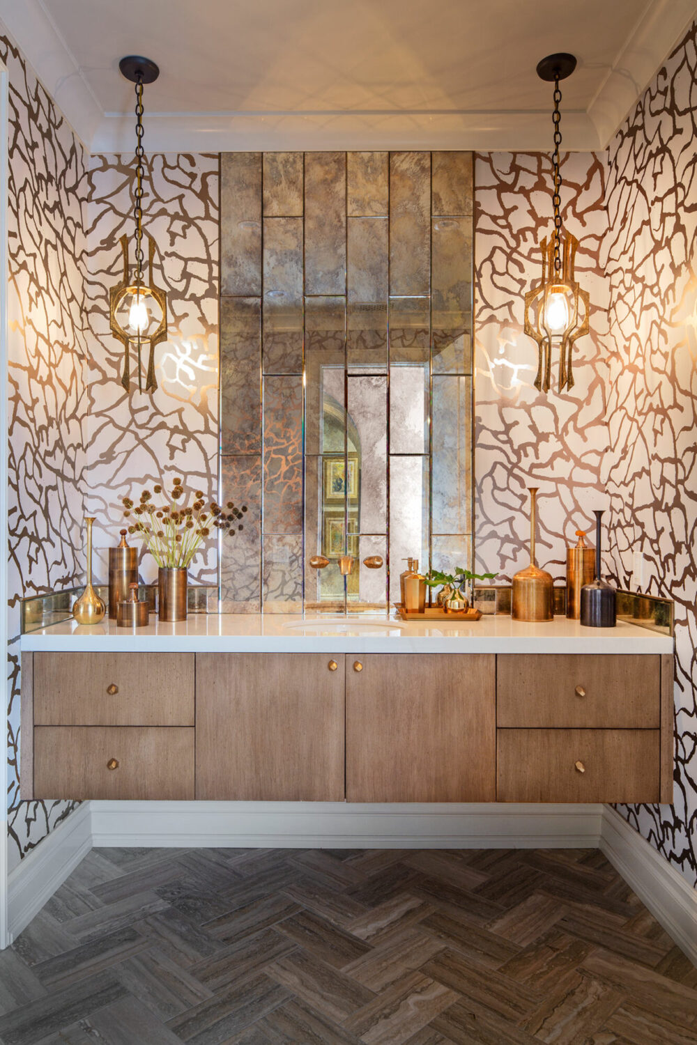 Bathroom with patterned wallpaper, mirrored backsplash, wood vanity, brass fixtures, decorative vases, and two pendant lights hanging from the ceiling.