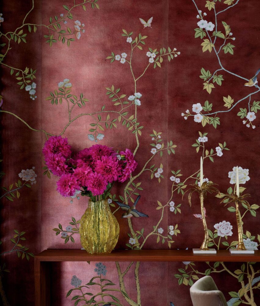A yellow vase with bright pink flowers sits on a wooden shelf against a red floral-patterned wallpaper, next to two gold candlesticks with white candles.