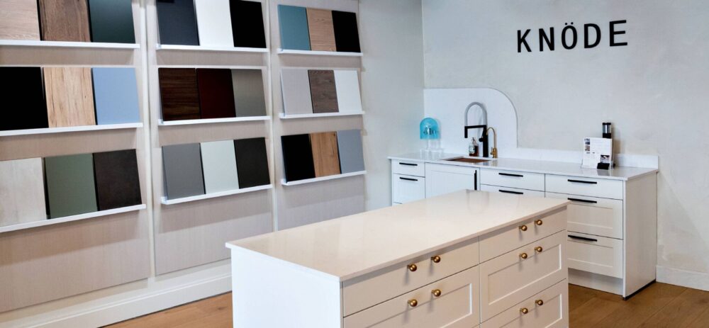 Modern showroom with a white island, display of various cabinet finishes on the wall, and a small sink area beneath the word “KNÖDE” on the wall.