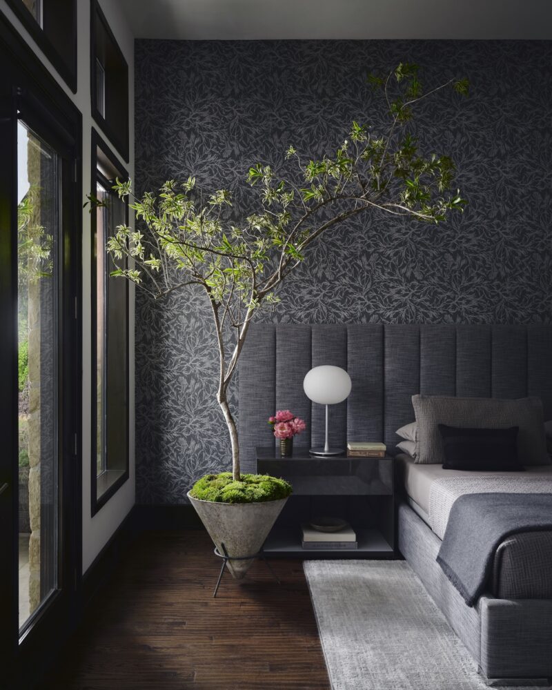 Modern bedroom with dark floral wallpaper, a large potted tree next to the bed, grey bedding, and a bedside table with a lamp and pink flowers. Natural light enters from glass doors.