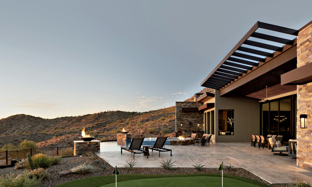 Modern patio with lounge chairs and fire pits overlooking desert hills and a putting green. Warm tones, open sky; serene and inviting atmosphere.