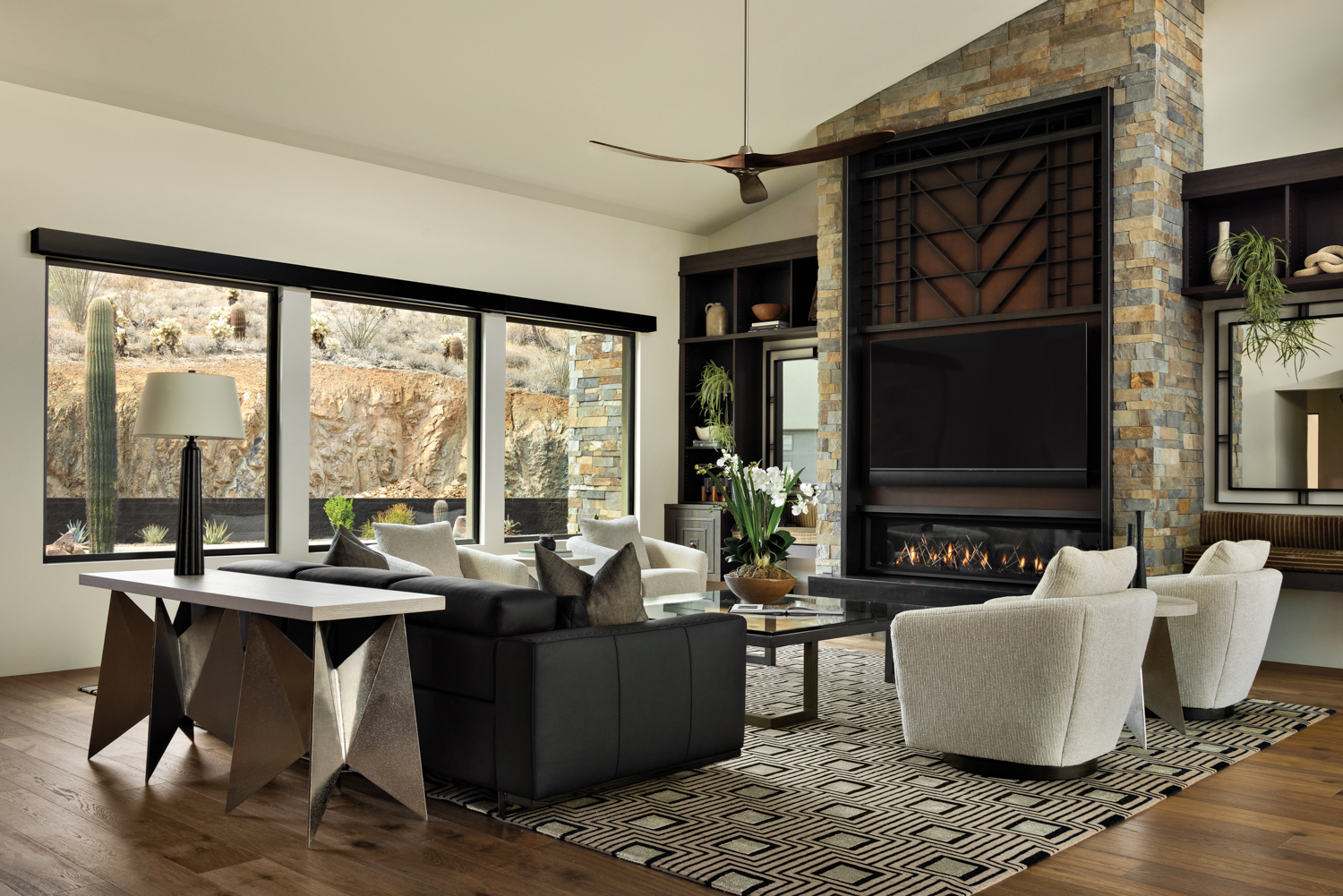 Modern living room with large windows showcasing a desert view. Features include a stone fireplace, black and white furniture, and a geometric rug. Cozy and stylish ambiance.