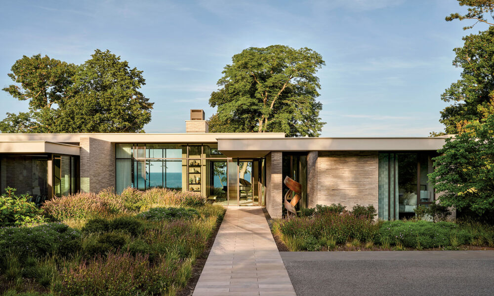 Modern single-story house with large windows, surrounded by lush greenery and trees. A stone path leads to the entrance, conveying a serene atmosphere.