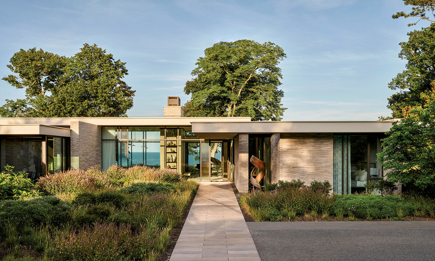 Modern single-story house with large windows, surrounded by lush greenery and trees. A stone path leads to the entrance, conveying a serene atmosphere.