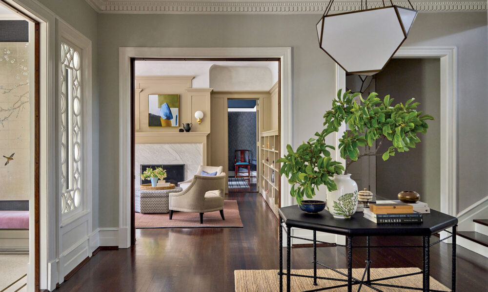 Bright, elegant hallway with dark wood floors and greenery on a black console table, leading to a living room with a fireplace, art, and neutral furnishings.