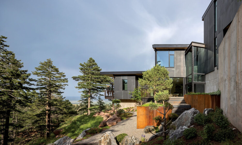 Modern house with large windows and concrete exterior, surrounded by pine trees and landscaped rocks on a hillside under a partly cloudy sky.