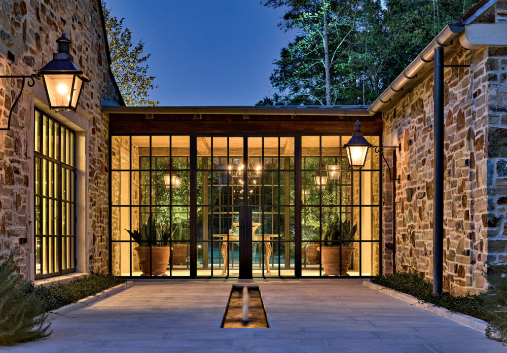 Elegant stone courtyard at dusk with glowing lanterns and large glass doors. Inside, warm lighting and potted plants create a cozy, inviting atmosphere.