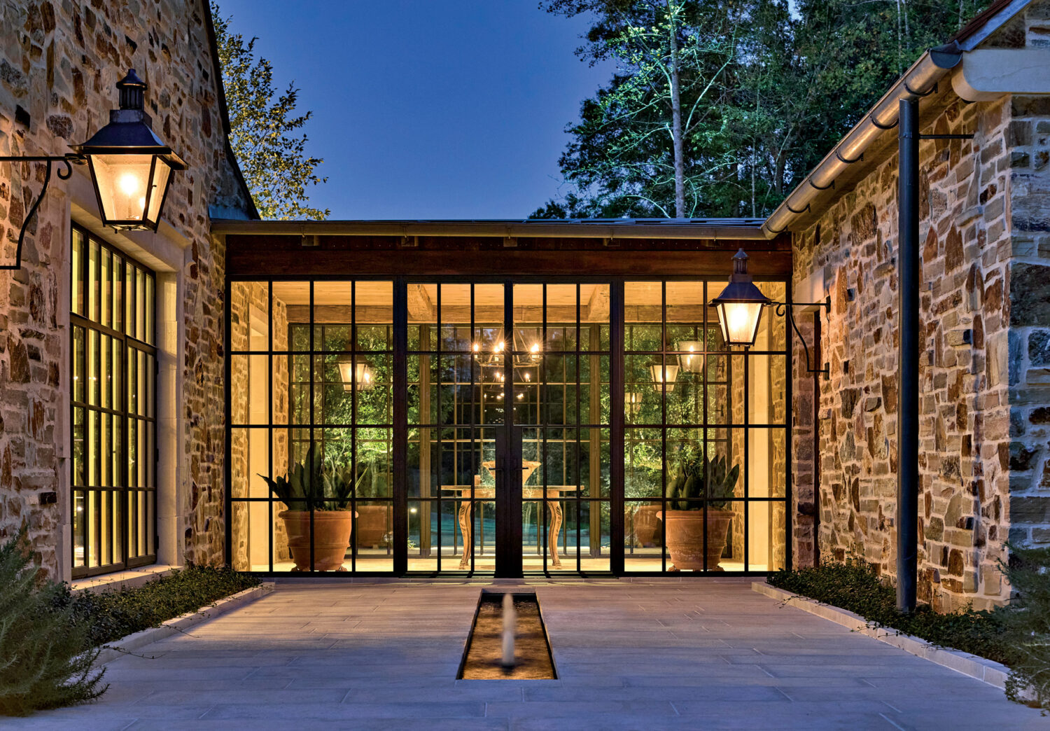Elegant stone courtyard at dusk with glowing lanterns and large glass doors. Inside, warm lighting and potted plants create a cozy, inviting atmosphere.