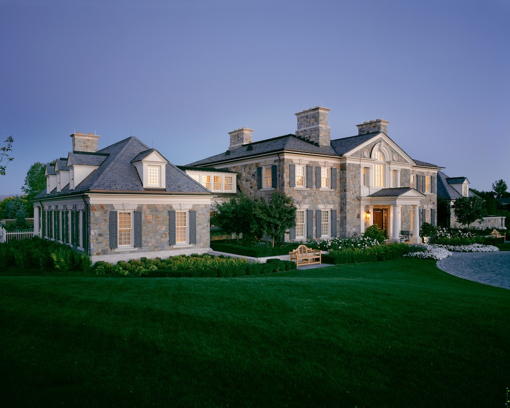 Large stone mansion with multiple chimneys, lit windows, manicured lawn, and a driveway, photographed at dusk.