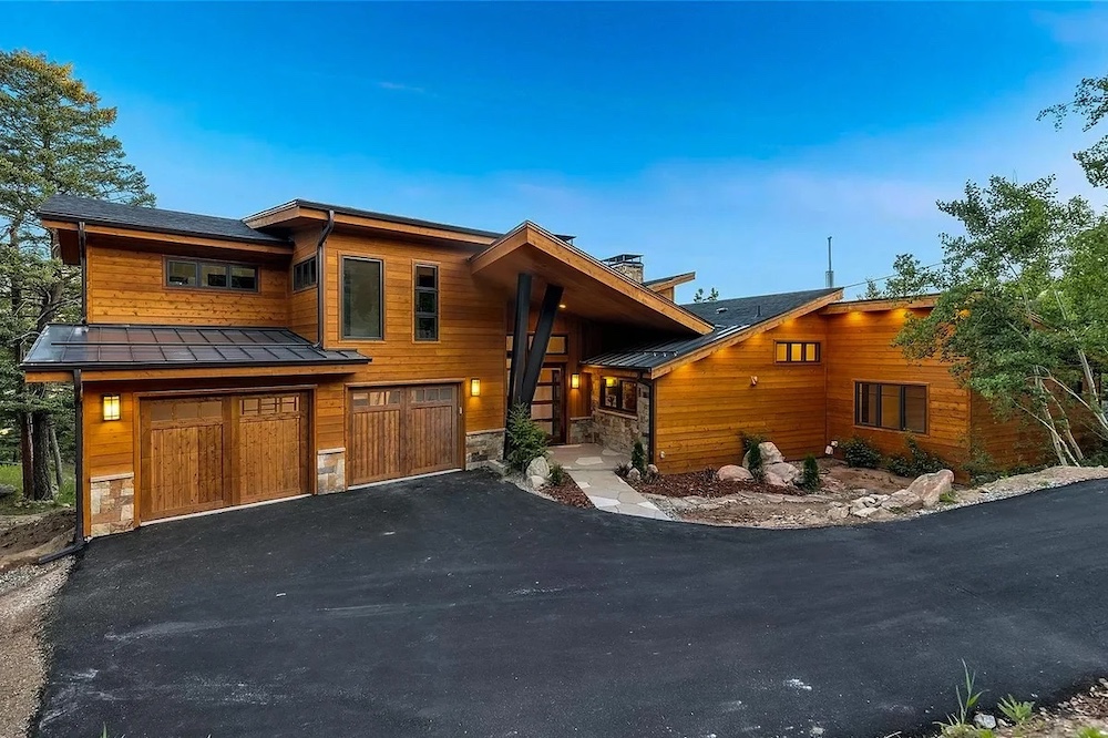 Modern wood-sided house with large windows, two-car garage, and angular roofline, surrounded by trees and a paved driveway at dusk.