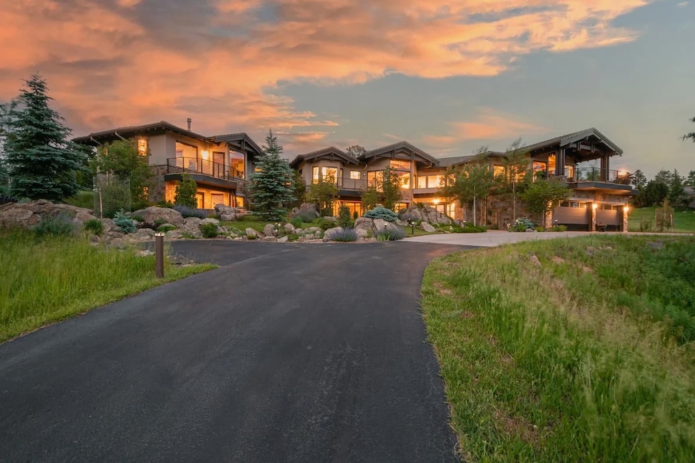 Modern multi-level house with large windows and warm interior lighting, set amid landscaped grounds and greenery, with a paved driveway leading up to the entrance at sunset.