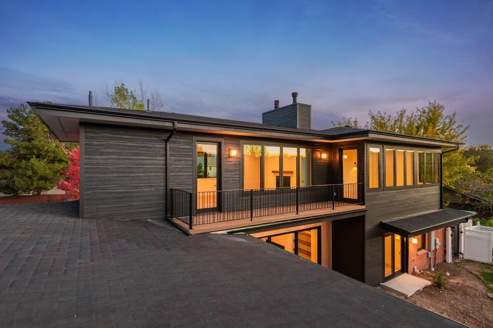 Modern two-story house with dark wood siding, large windows, and a wraparound balcony, photographed at dusk with interior lights on.