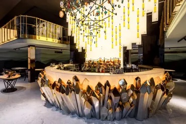 A circular bar with a metallic, crystal-like base and a chandelier of hanging bottles, surrounded by seating in a modern, upscale interior.