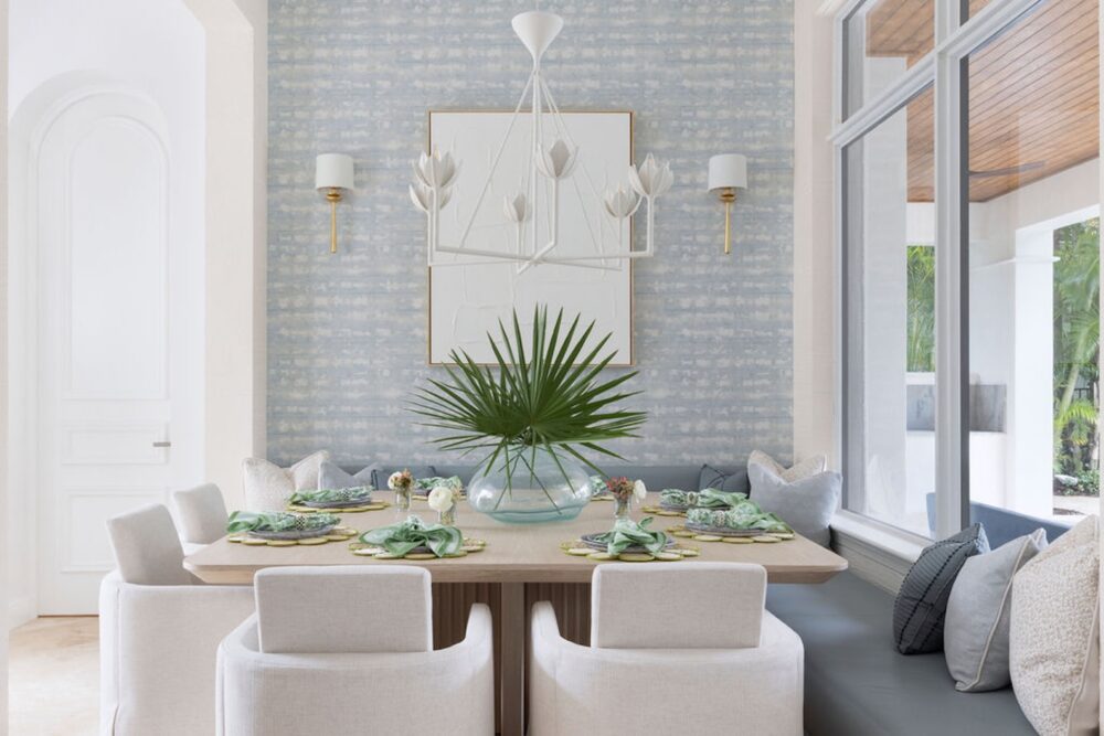 A modern dining room with a light wooden table, six white chairs, a green leafy centerpiece, and a geometric chandelier. Large windows and patterned wallpaper are visible.