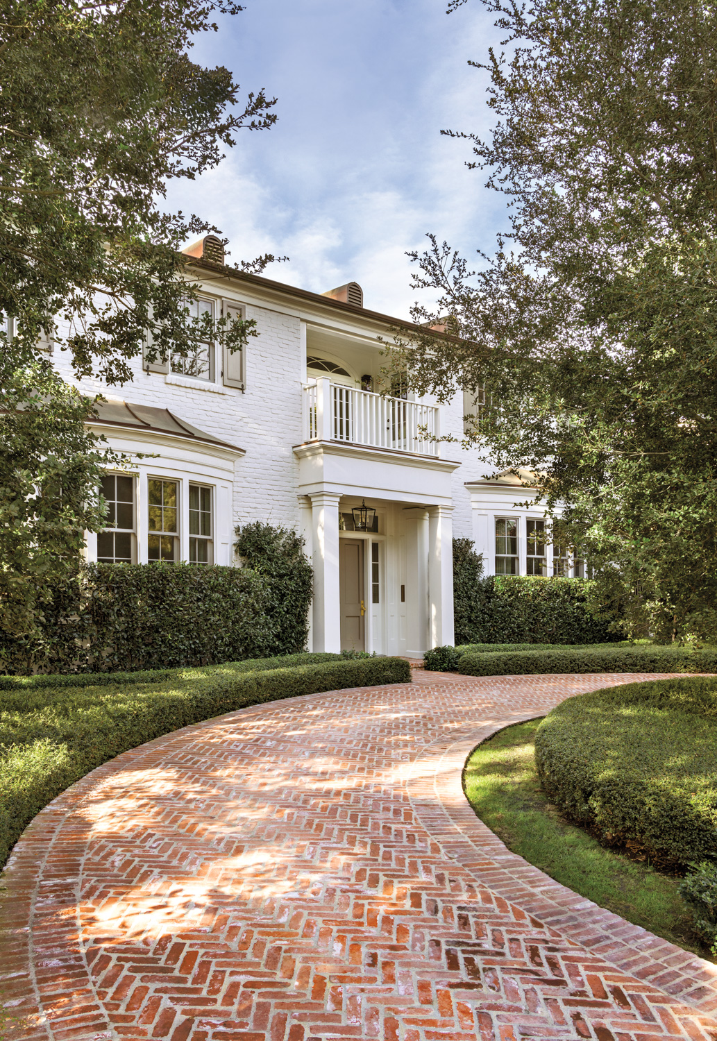 Elegant white two-story house with a front balcony, surrounded by lush greenery. Red brick pathway curves toward the entrance, creating a welcoming ambiance.