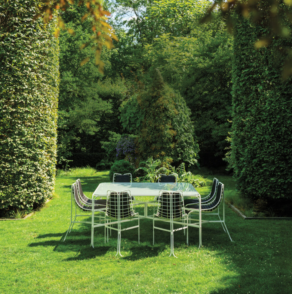 A metal outdoor dining table with eight chairs is set on a green lawn, surrounded by tall, manicured hedges and lush trees in a garden.