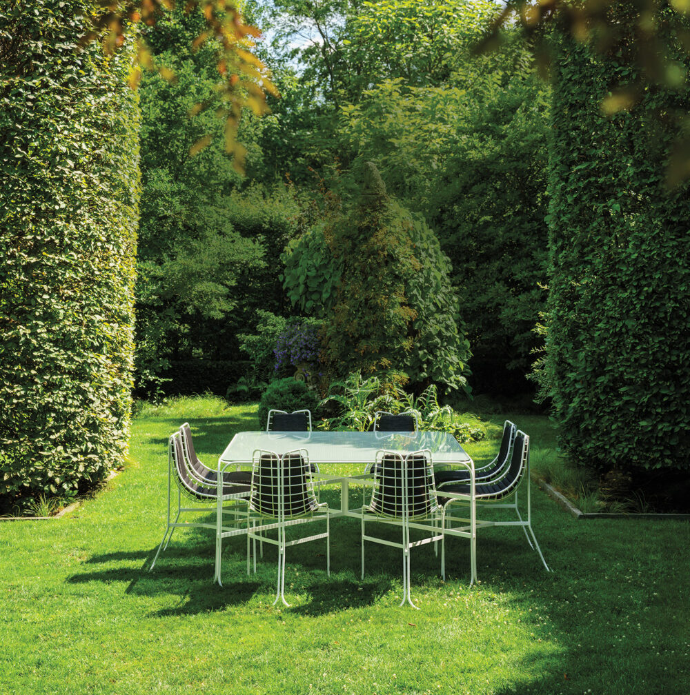 A metal outdoor dining table with eight chairs is set on a green lawn, surrounded by tall, manicured hedges and lush trees in a garden.